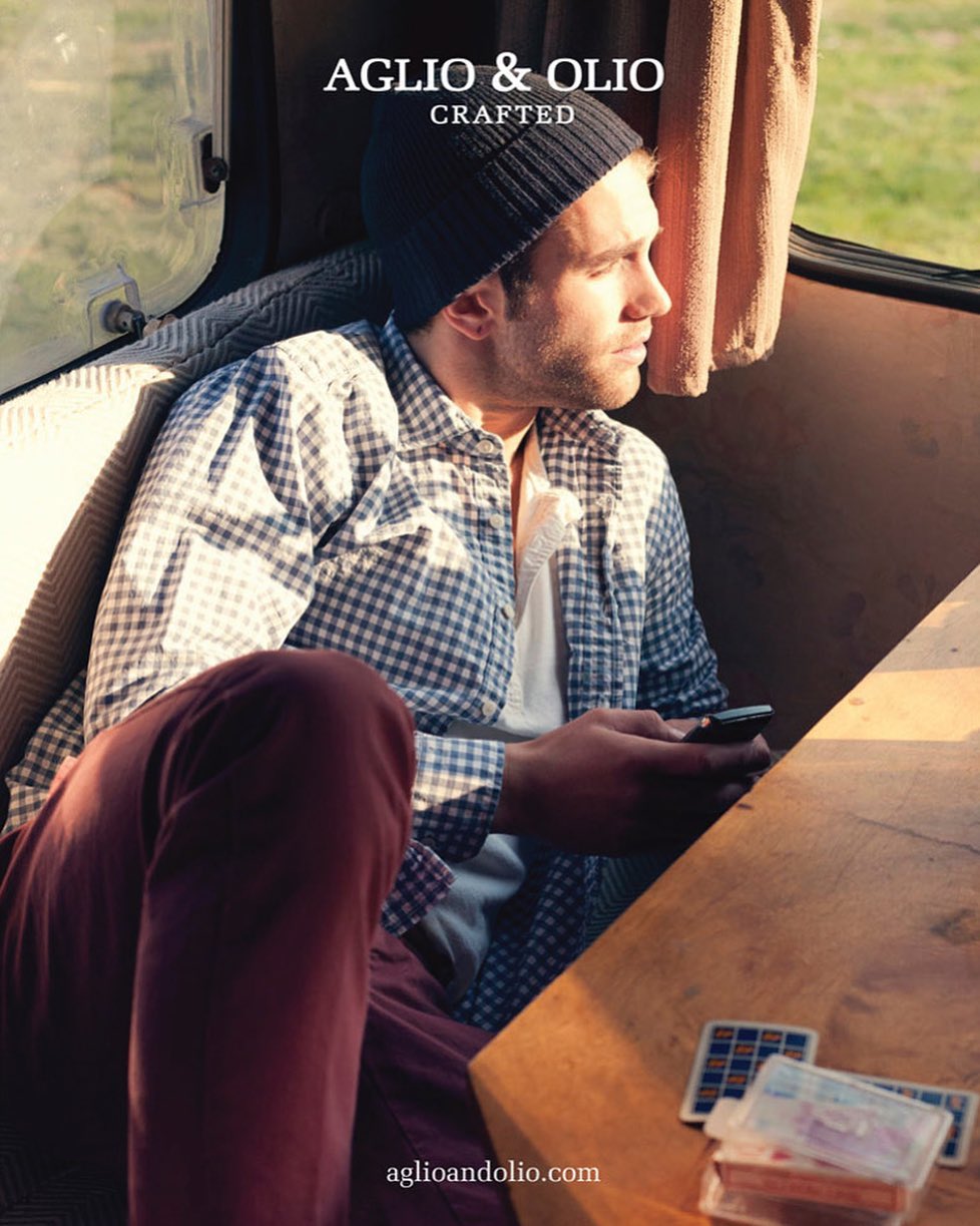 Man in flannel shirt and beanie sits in van, looking out window with phone in hand. Wooden table with playing cards nearby.