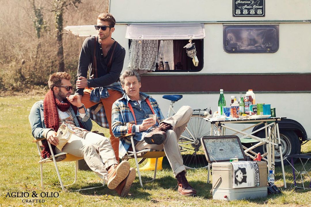 Three adult men are seated in chairs beside a vintage RV in an outdoor grassy field, enjoying drinks and relaxing on a sunny day.