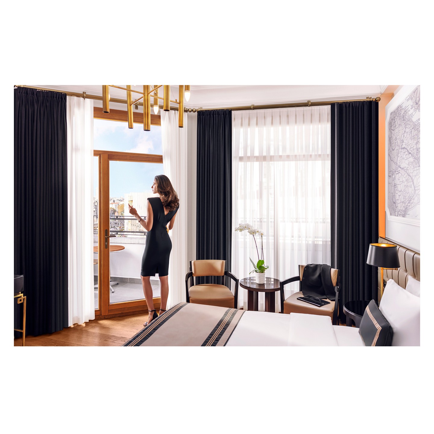 Woman in black dress on hotel balcony with drink, city view. Modern room features gold light fixture and stylish furniture.