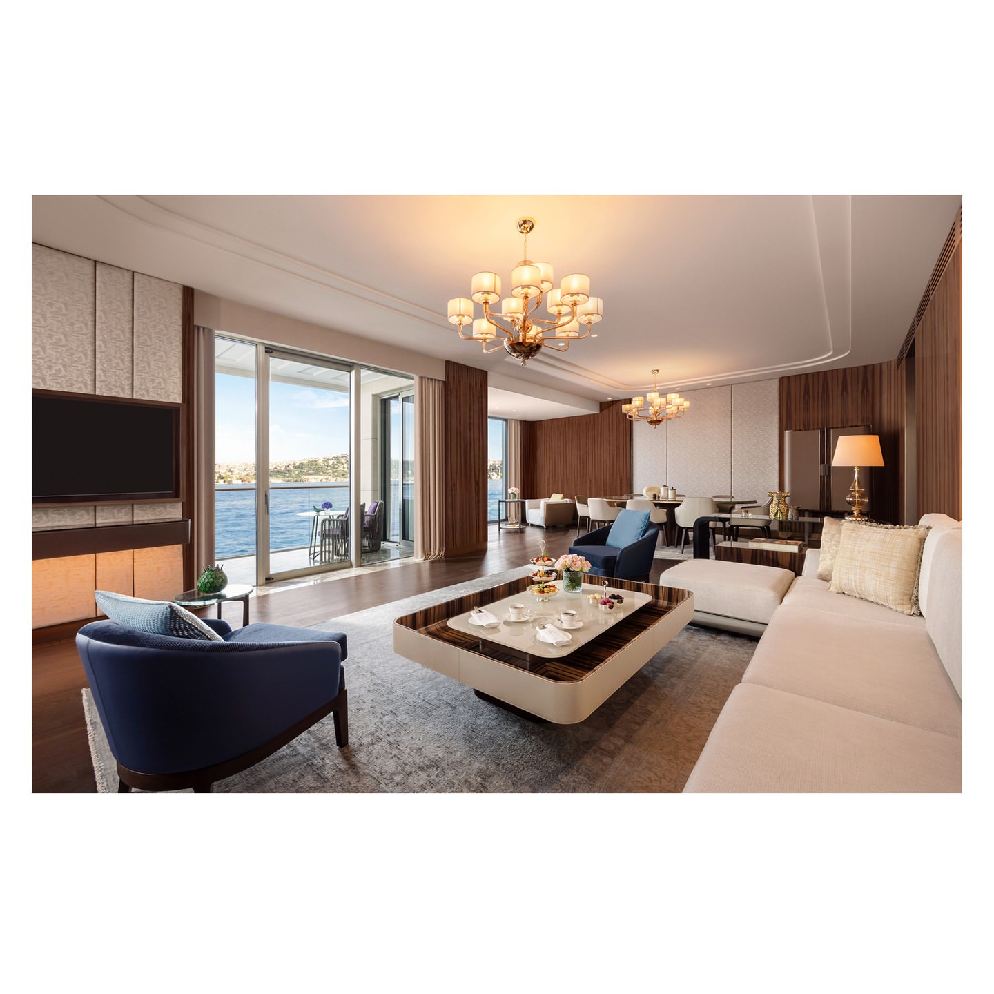 Opulent living room interior with a beige sectional sofa, blue accent chair, coffee table, chandelier, and a waterfront view.