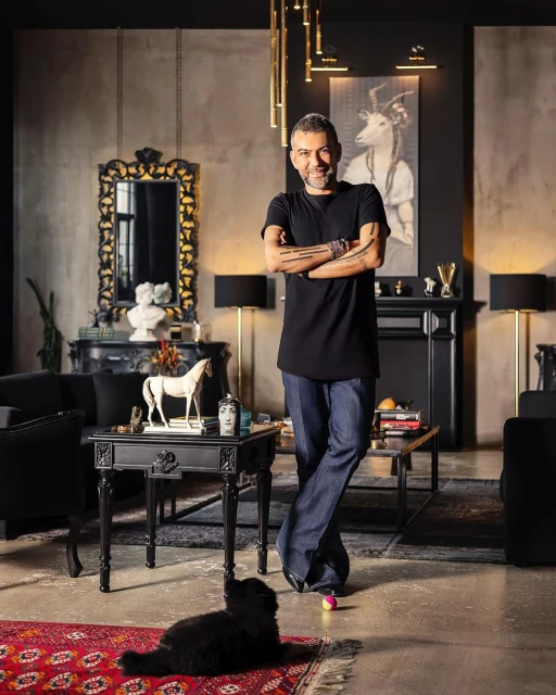 Man in a black shirt standing in a modernly furnished living room next to a black dog lying on a red patterned rug.
