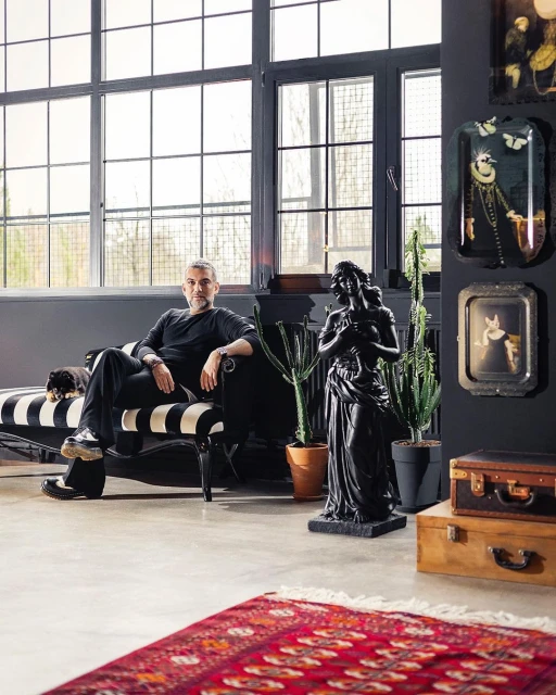 Stylish man in a black outfit lounges on a striped chaise, indoors, with modern decor, including a statue, plants, and decorative trays.