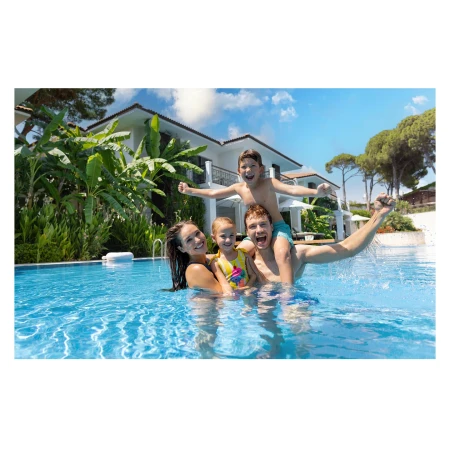 A happy family of four playing in a swimming pool outside a modern house with lush tropical plants and a bright blue sky.
