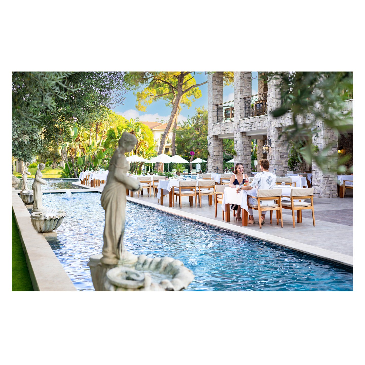 Outdoor restaurant scene features a woman and man seated at a table beside a pool with statues, set against a lush, green backdrop.
