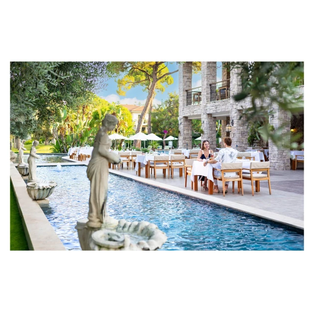 Outdoor restaurant scene features a woman and man seated at a table beside a pool with statues, set against a lush, green backdrop.