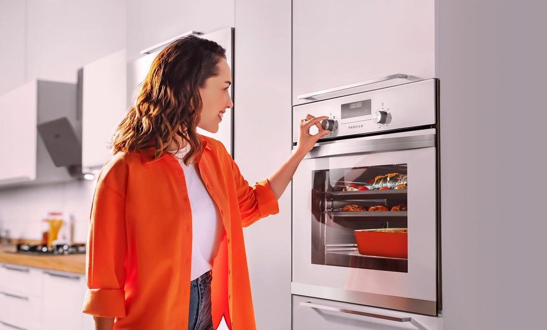 A woman in an orange shirt adjusts the settings on a stainless steel oven, with food visible inside, in a modern kitchen setting.