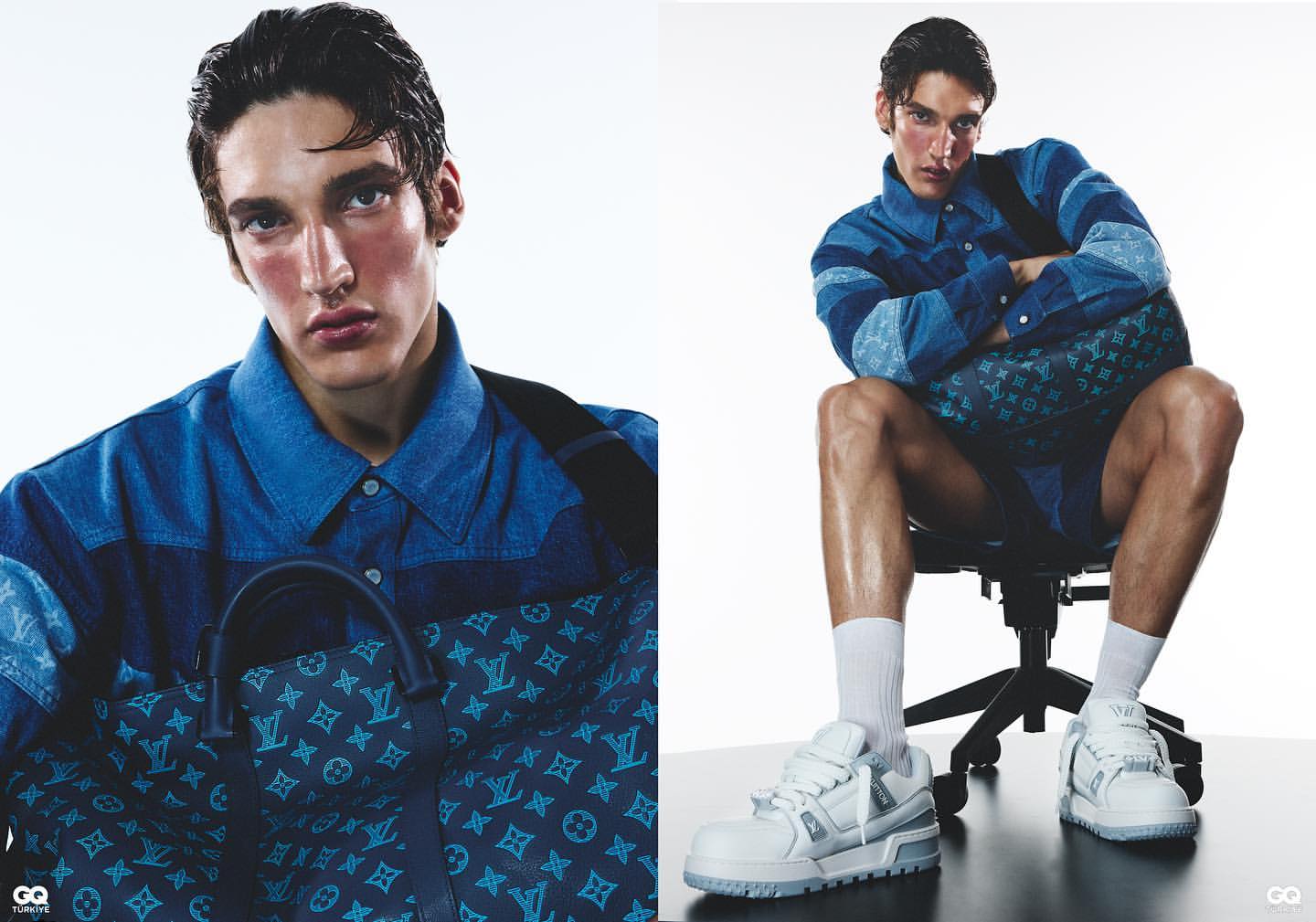 Male model in a patchwork denim shirt and Louis Vuitton bag, seated indoors with white sneakers in a fashion editorial shoot.