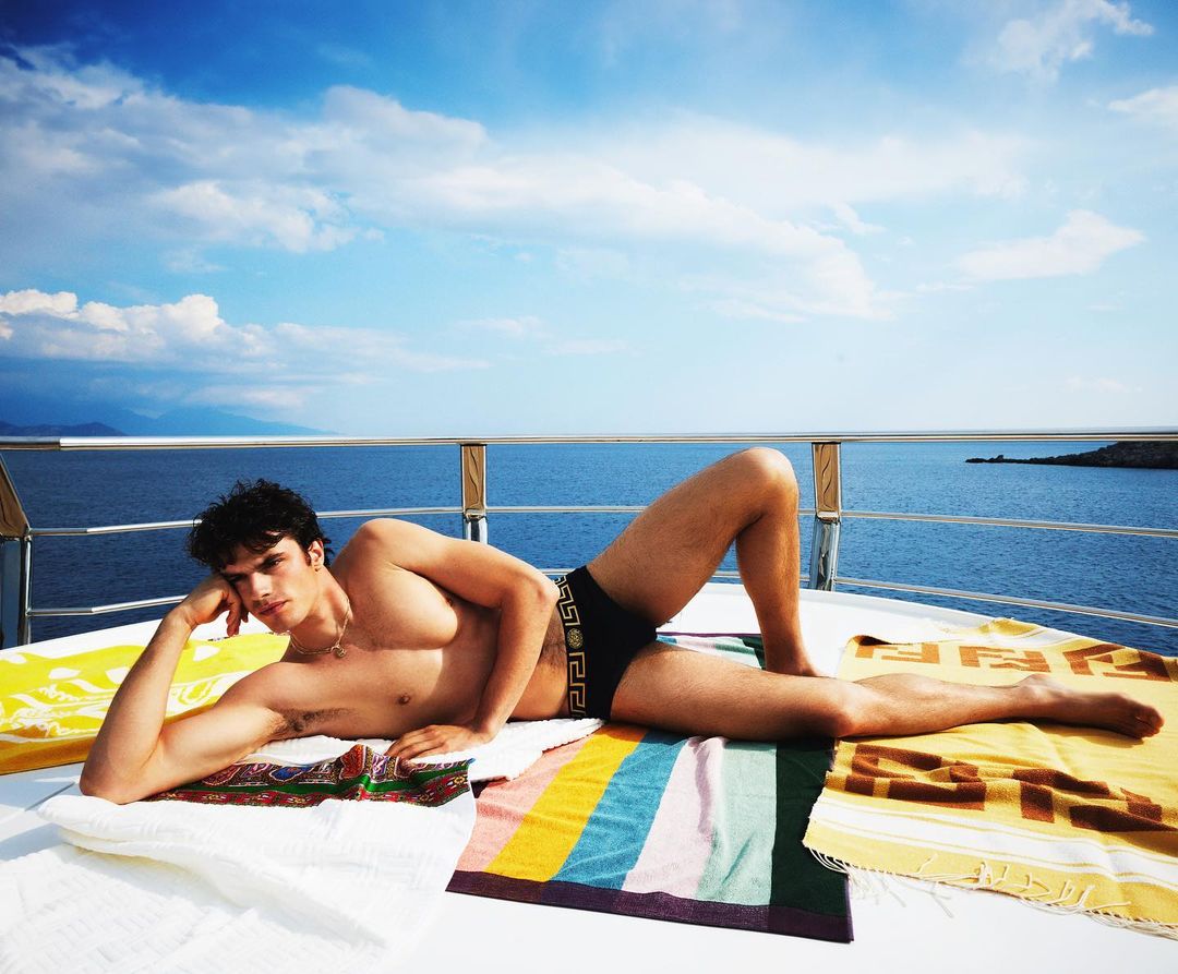 A tanned man with dark hair lounges on a boat deck near the ocean, wearing Versace swim trunks and laying on patterned towels on a sunny day.