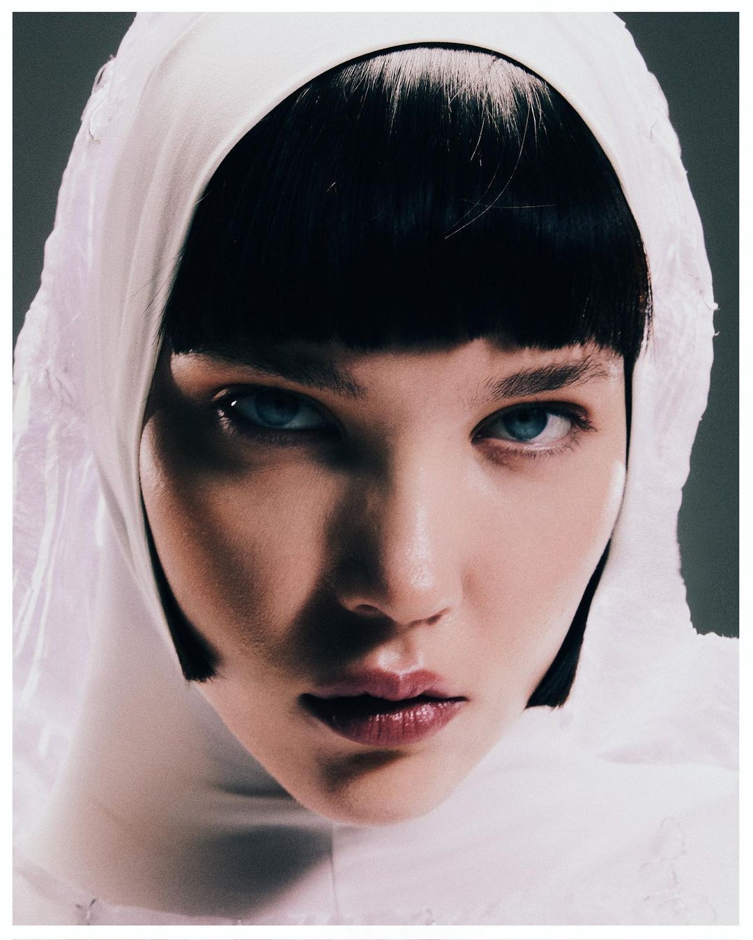 Close-up portrait of a young adult female with light skin, short black hair, blue eyes, and a white embroidered hood.