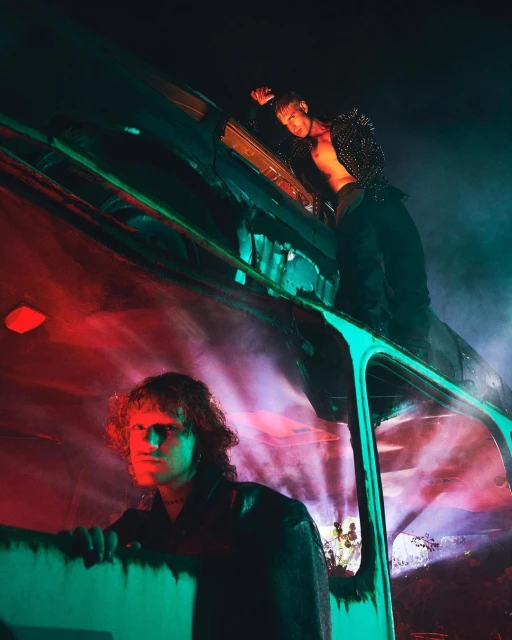 Two men pose in front of an old bus under red and teal lighting. One man stands on the roof, the other looks out the window.