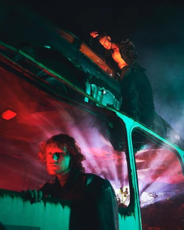 Two men pose in front of an old bus under red and teal lighting. One man stands on the roof, the other looks out the window.