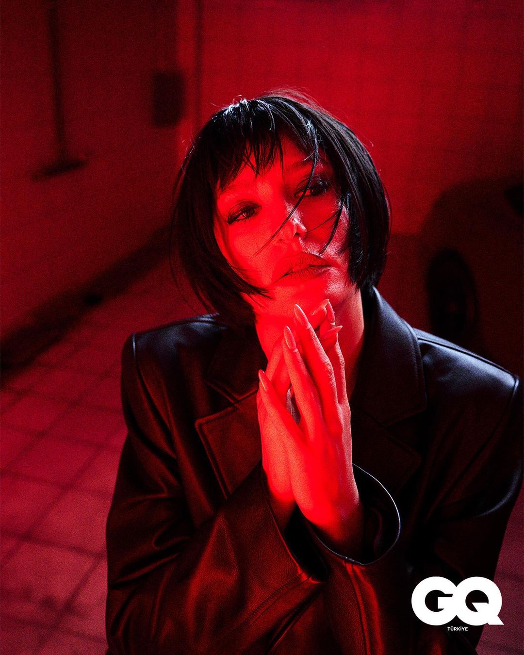 Fashion portrait of a young adult woman with a bob haircut, wearing a black leather jacket, hands clasped in a red-lit interior.