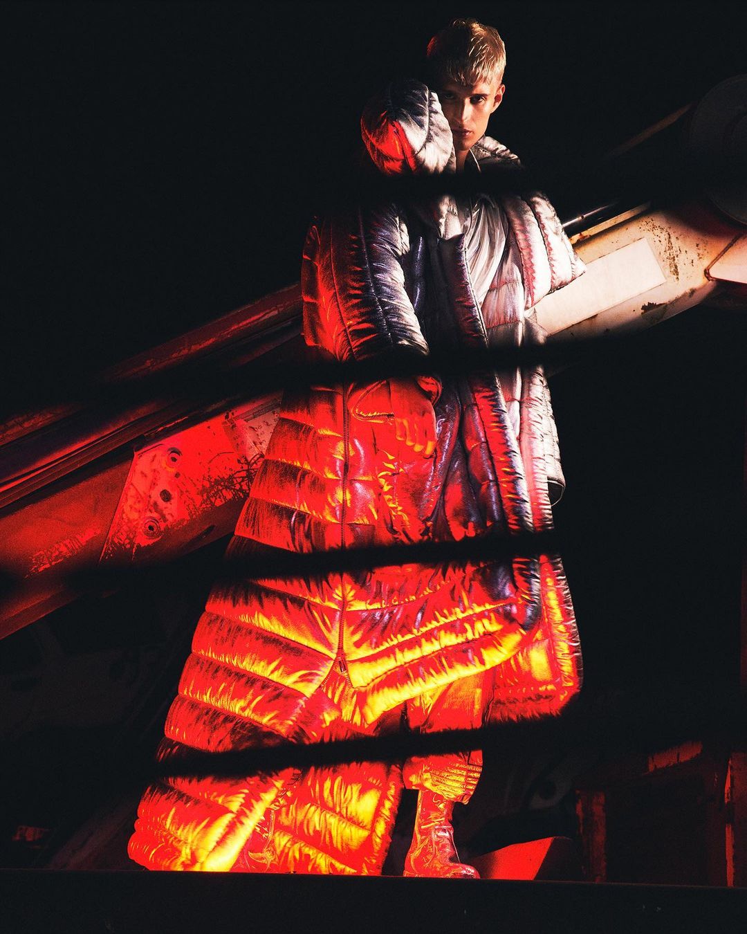 Full-length shot of a blond adult male in a hooded silver puffer coat with red light, with dark background. Fashion or editorial style.