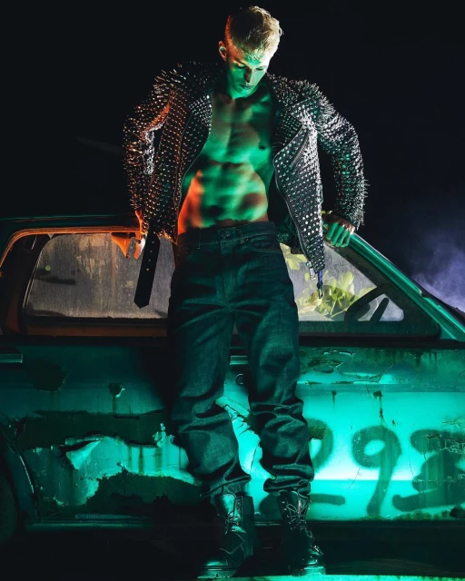 Male model poses with open studded jacket, jeans, and boots in front of a rusty car, illuminated with green and orange lighting.