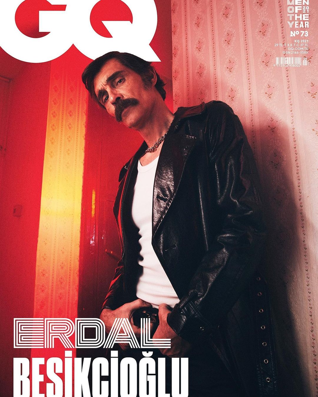 Man with a mustache wearing a black leather jacket and white t-shirt standing against patterned wallpaper.