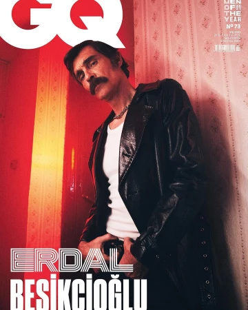 Man with a mustache wearing a black leather jacket and white t-shirt standing against patterned wallpaper.