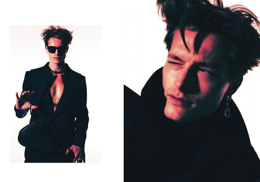Editorial shot of a stylish adult male in a black suit and sunglasses posing in a studio setting with red lighting, wearing jewelry.