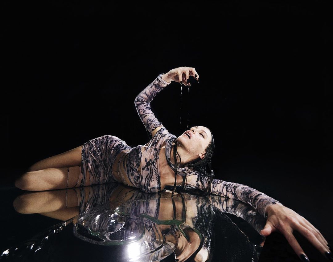 A woman with dark hair, lying on a reflective surface against a dark background, pours liquid down her face in a dramatic pose.