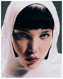 Close-up portrait of a woman with dark blunt bangs wearing a white head covering and dark eye makeup against a grey background