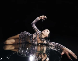 Model lying on reflective dark surface in printed long-sleeve dress letting water drip from raised hand against black backdrop.