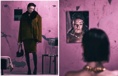 Model in olive green coat and skirt standing against a textured pink wall with a chair and a framed mirror reflection