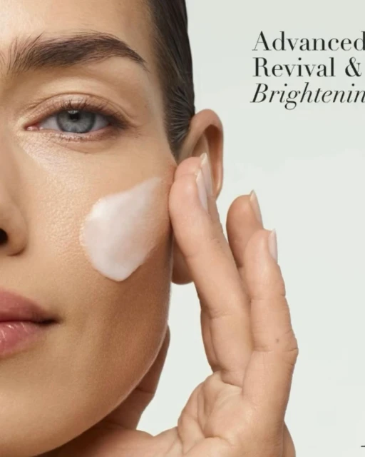 Close-up of a woman's face with light skin tone and blue eyes, applying white cream to her cheek, advertising beauty product.