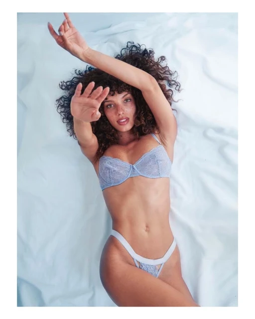 Woman in a blue lace lingerie set poses while lying on a white sheet in a brightly lit indoor setting. She is looking at the camera.