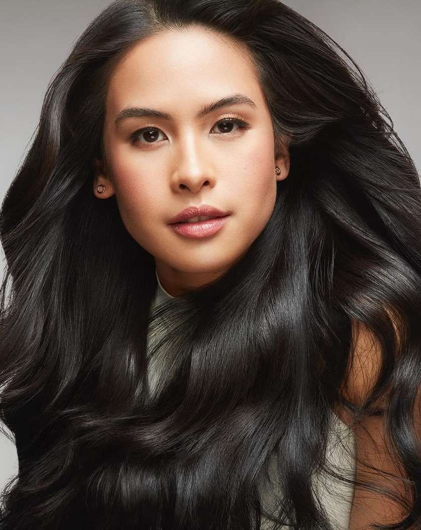 Close-up portrait of a woman with long dark wavy hair wearing a white top against a grey background by Erez Sabag.