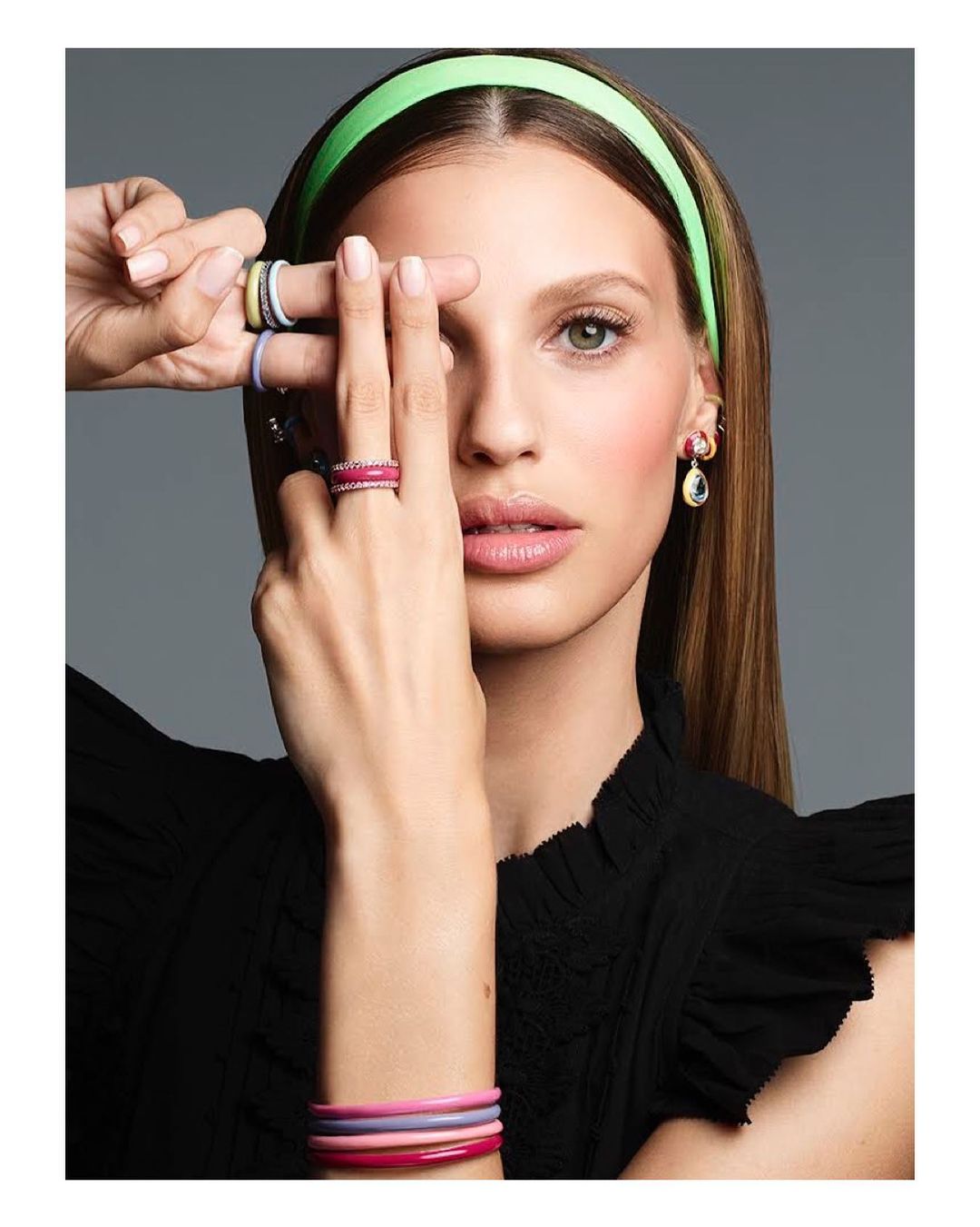 Close-up of a woman with blonde hair wearing a green headband and rings, posing in a black top against a grey background.