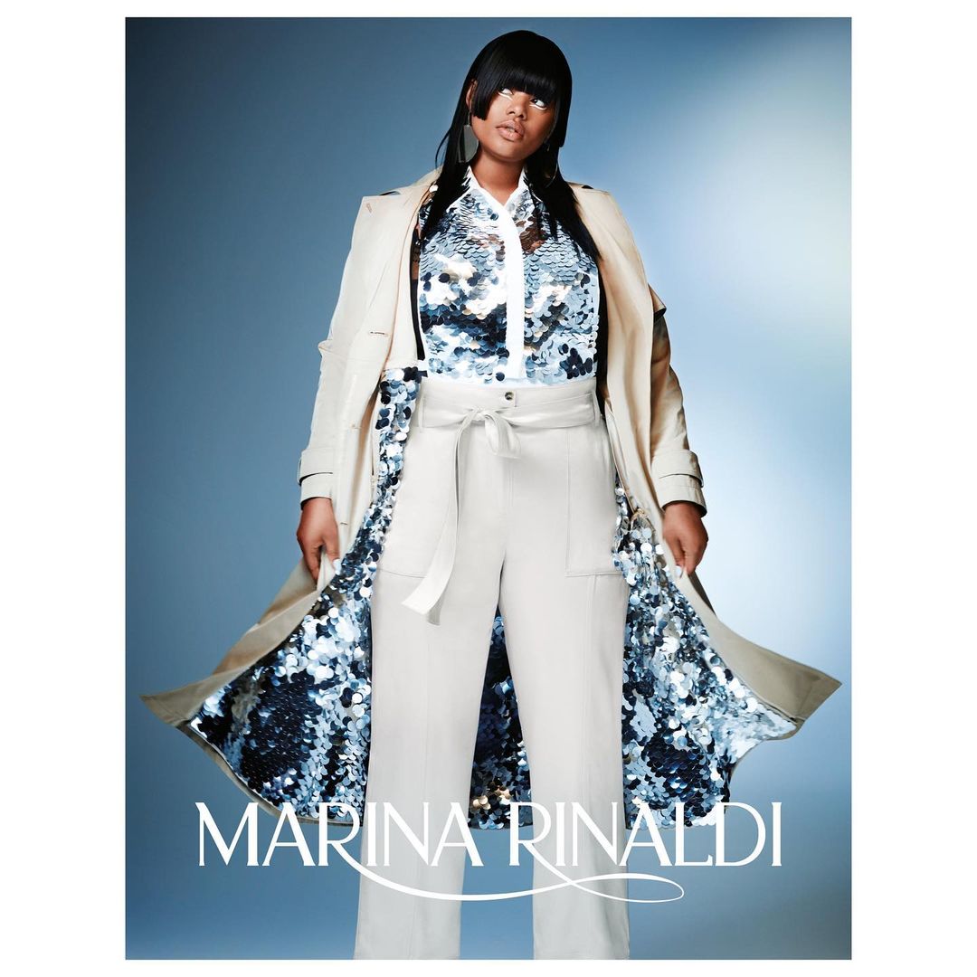Plus-size fashion model poses in a cream-colored belted coat and wide-leg trousers with a sequined blouse against a gradient blue backdrop.