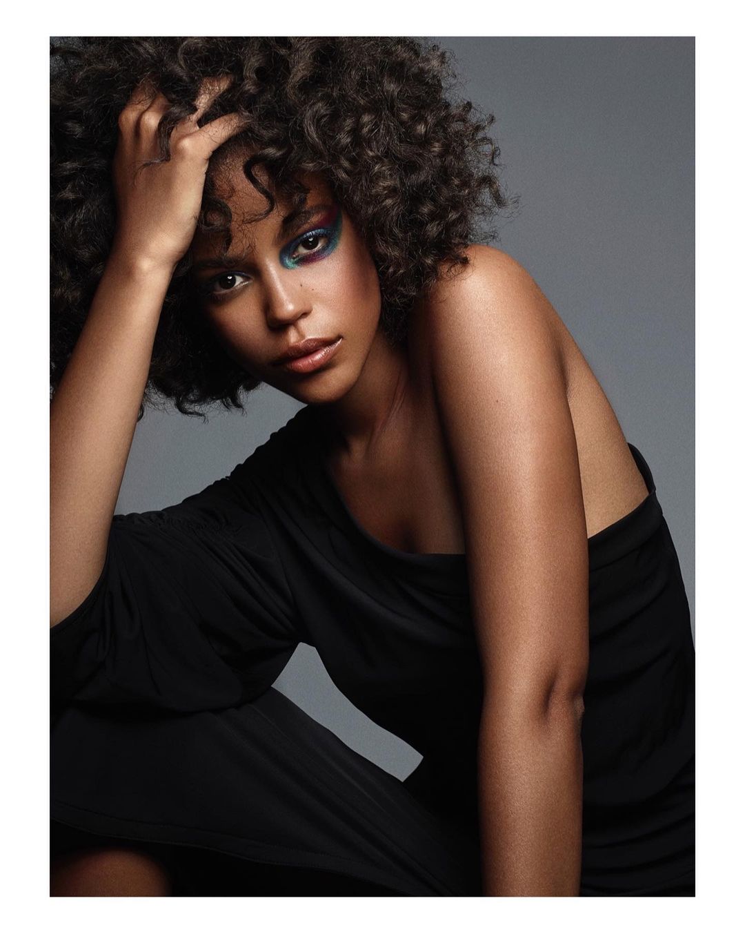 Close-up studio portrait of a Black woman with curly hair, wearing a black dress and colorful eye makeup, gazing directly at the camera.