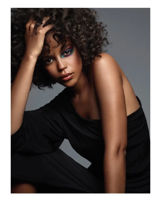 Close-up studio portrait of a Black woman with curly hair, wearing a black dress and colorful eye makeup, gazing directly at the camera.