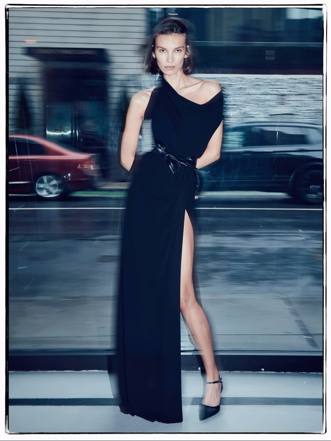 Elegant woman poses in a long black dress with a slit, accessorized with a belt and heels, against a blurred city street backdrop.