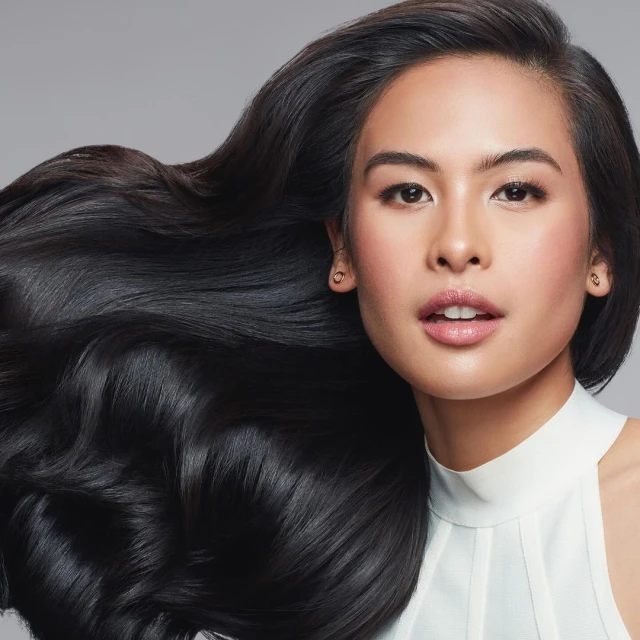 Brunette woman with long dark hair blowing in the wind, wearing a white top, posing in a beauty shot against a gray background.