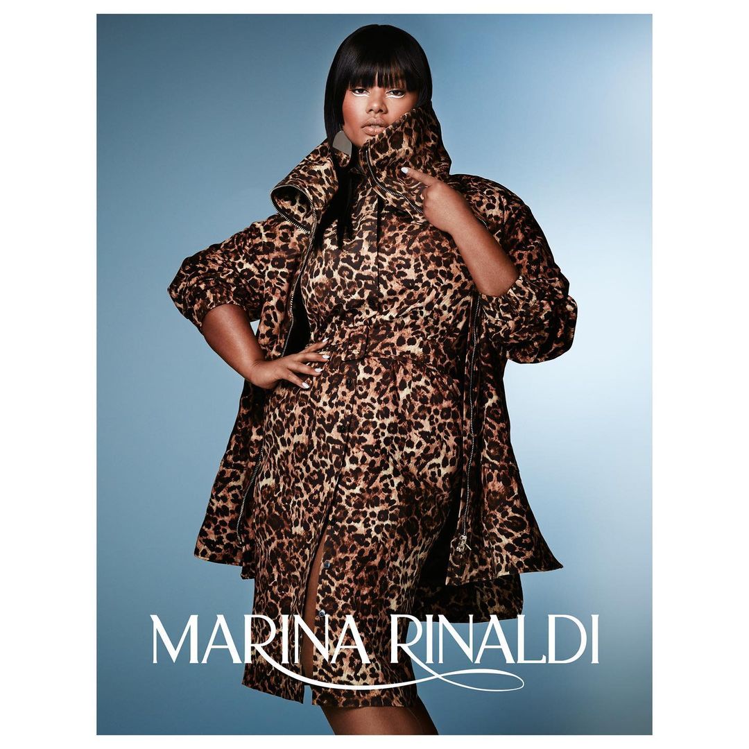 Plus-size woman in leopard-print dress and coat poses against blue backdrop; fashion-forward and confident in the photo studio.