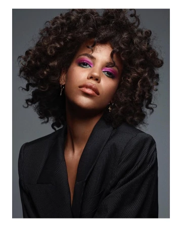 Portrait of a woman with voluminous dark curly hair, vibrant pink eyeshadow, and a black blazer against a grey background.