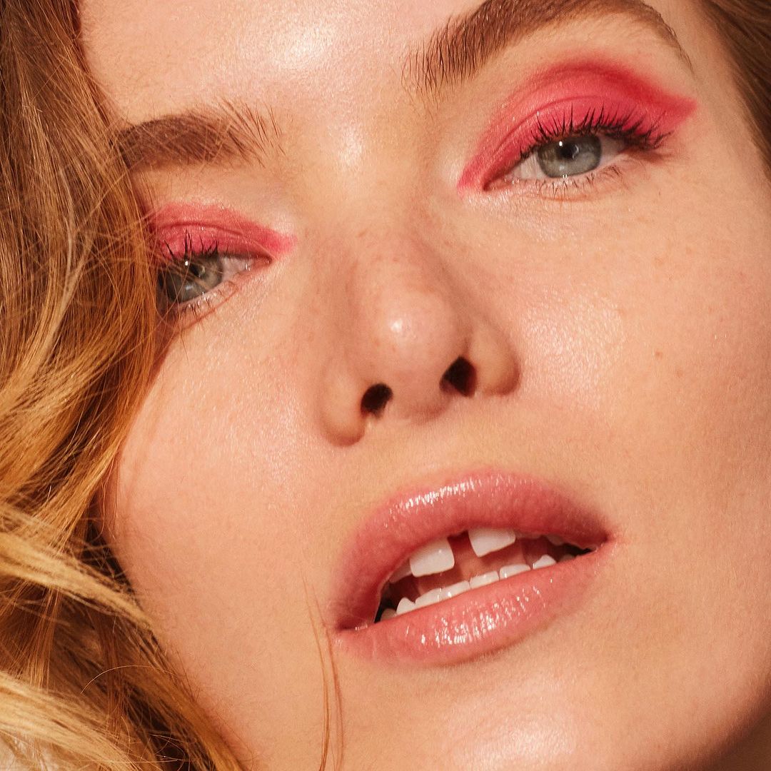 Close-up shot of a young woman with light skin, blue eyes, and reddish blonde hair, wearing pink eyeshadow. Beauty shot.