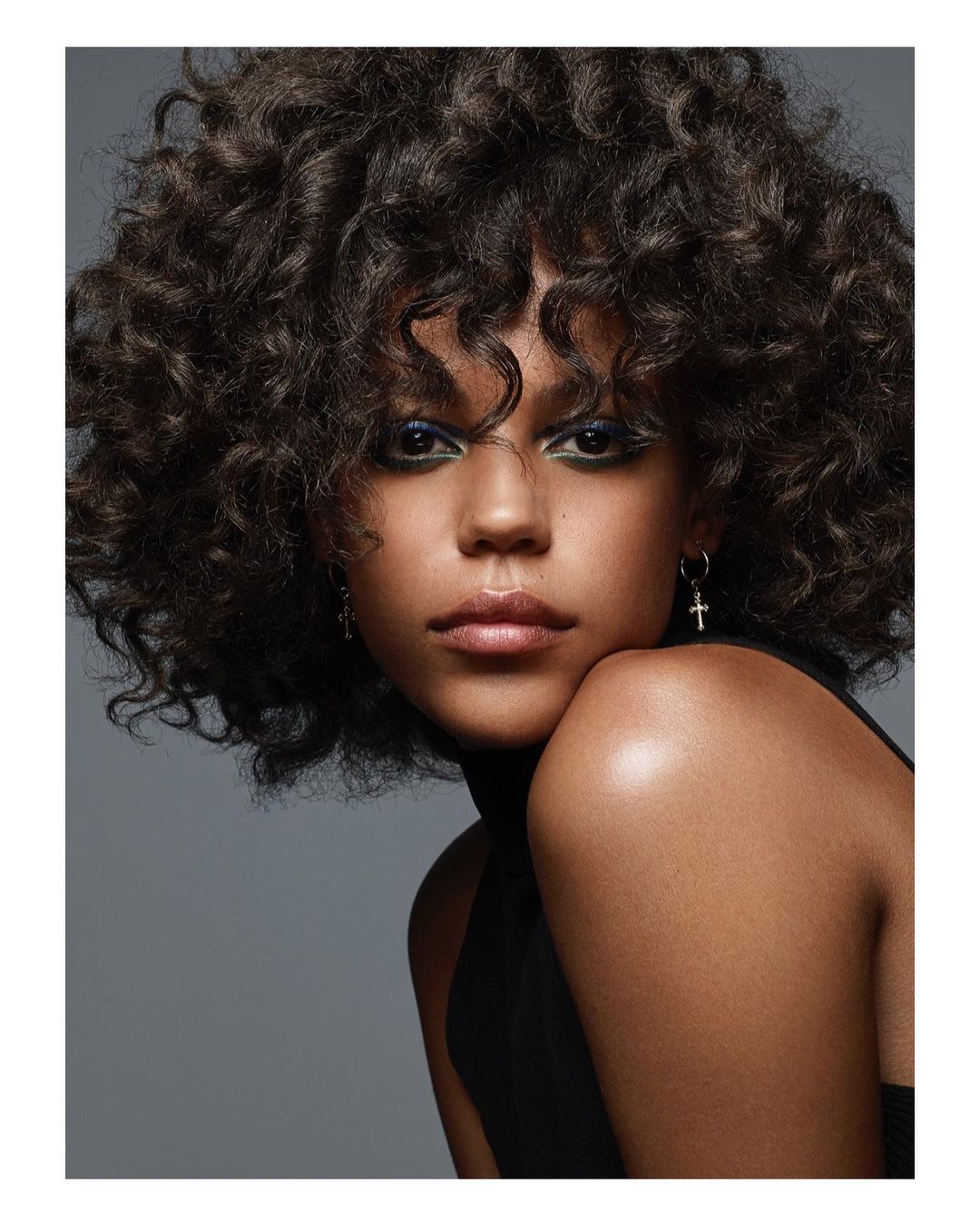 Portrait of a young adult female with dark, curly hair and green eyeshadow, wearing a black top against a grey backdrop.