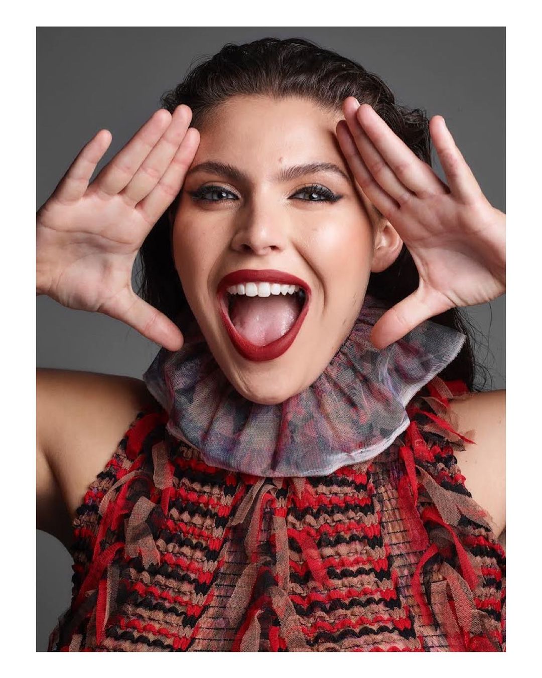 Excited adult female poses in red patterned top with dramatic ruffled collar, hands framing face, large smile, against grey backdrop.