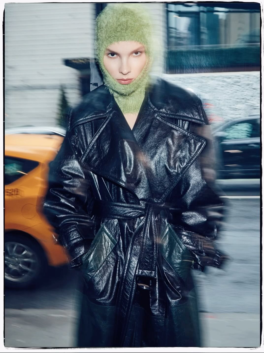 A woman in a black patent leather trench coat and green balaclava stands outside on a city street with a blurred taxi in the background.