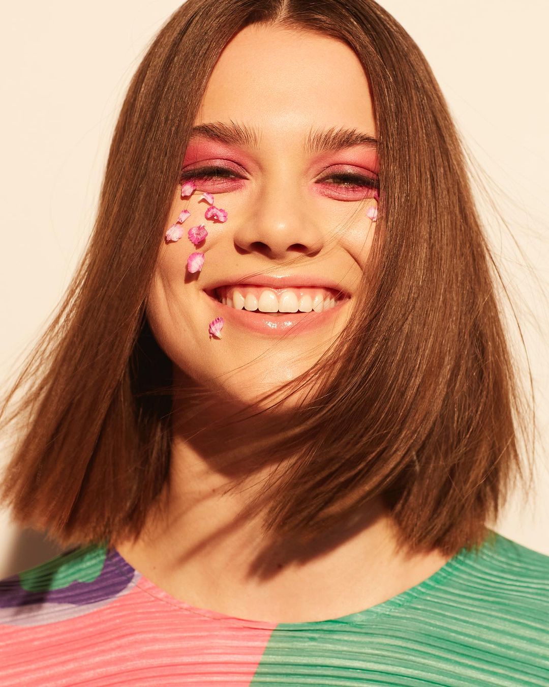 Close-up of a smiling young woman with short brown hair and pink makeup, adorned with small pink petals on her face.