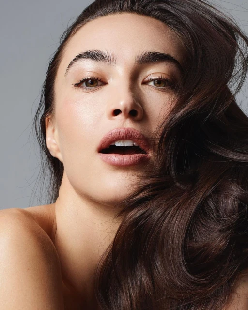 Close-up shot of a woman with long, wavy brown hair and natural makeup, looking directly at the camera against a neutral grey backdrop.