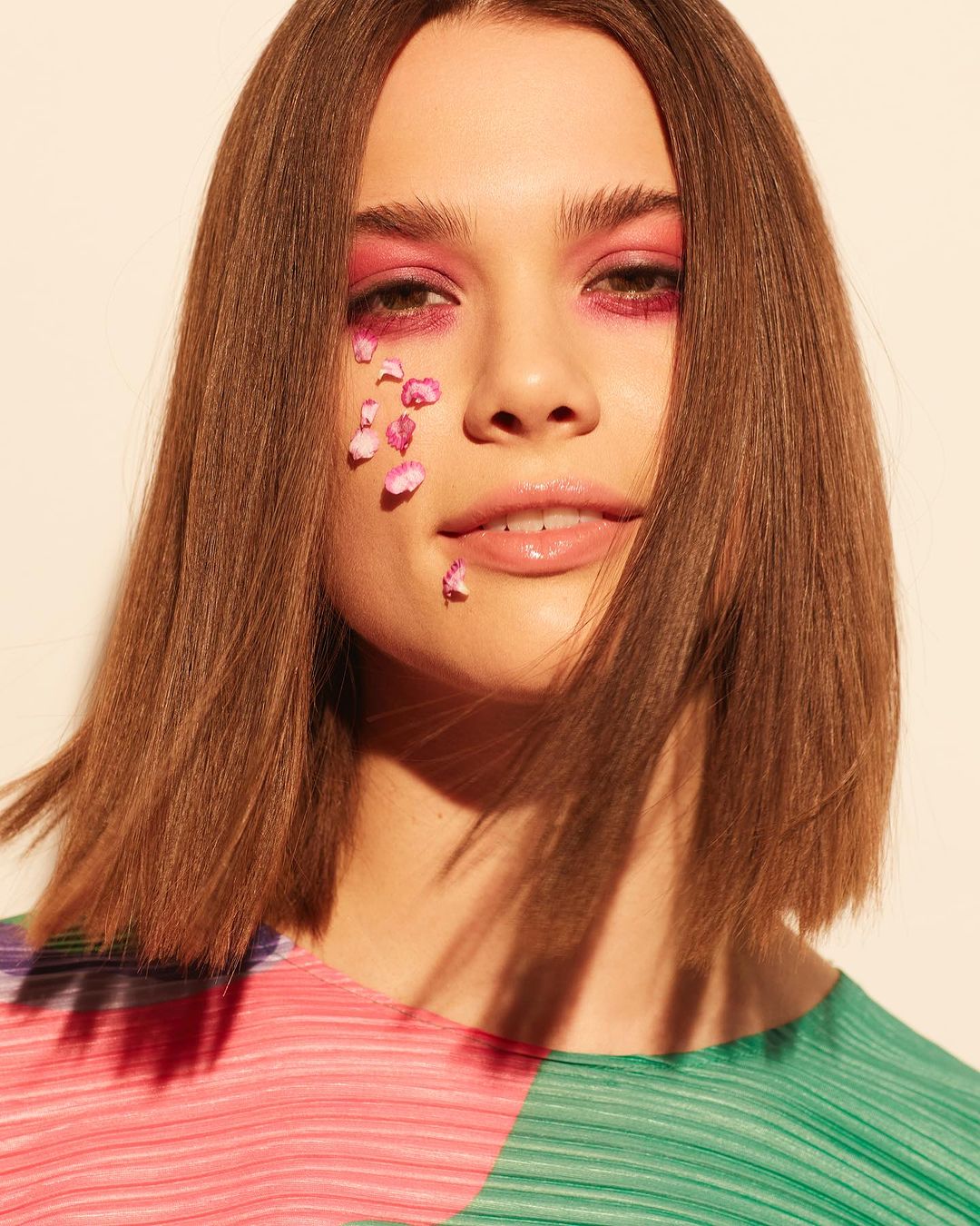 Beauty portrait of a brown-haired woman with pink eyeshadow and flower petals on her face against a cream-colored backdrop. Beauty shot.