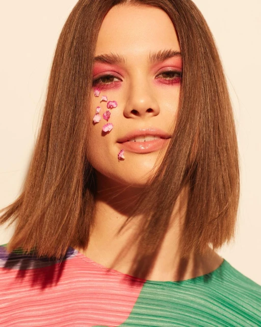 Beauty portrait of a brown-haired woman with pink eyeshadow and flower petals on her face against a cream-colored backdrop. Beauty shot.