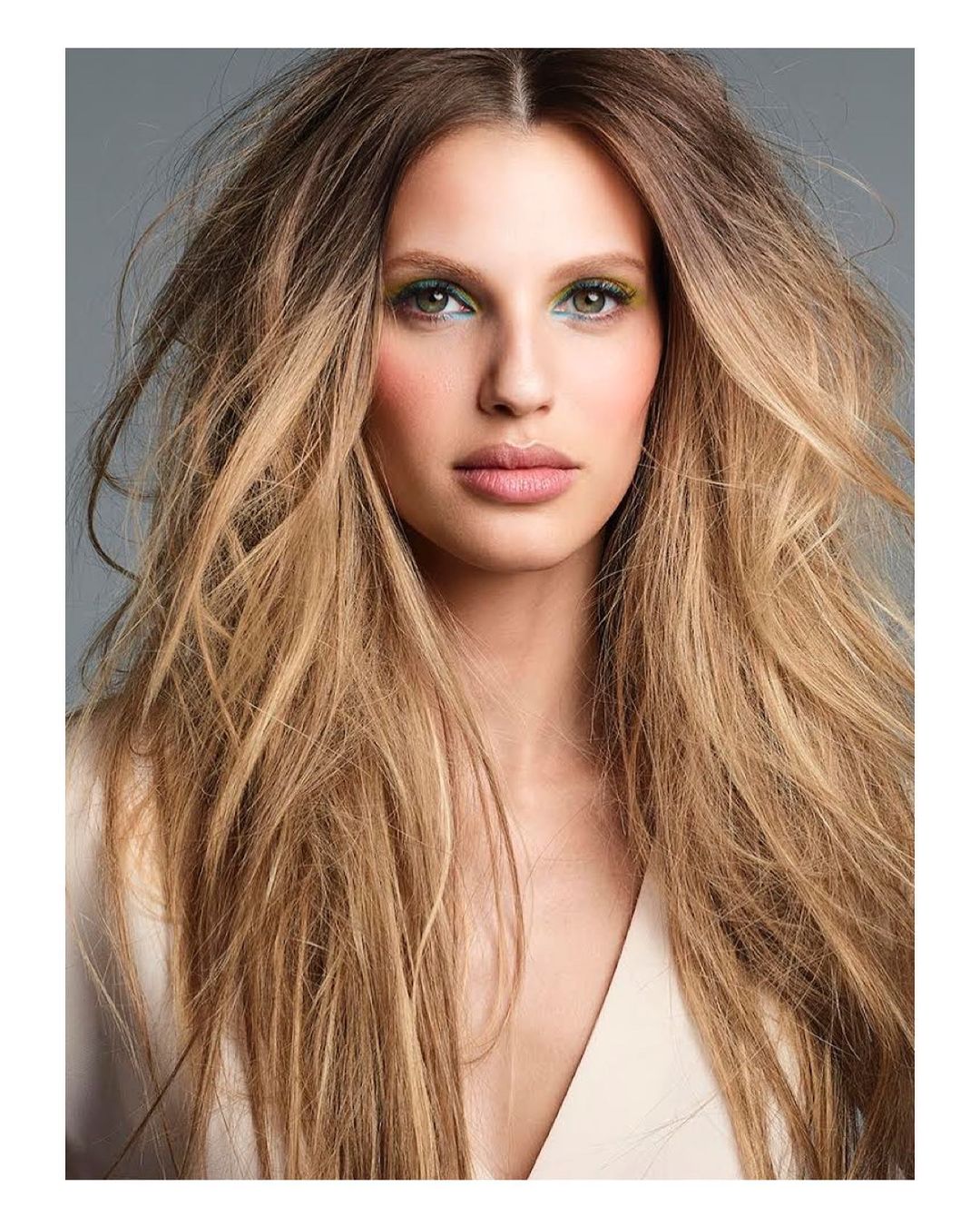 Close-up shot of a woman with long blonde hair, light skin and green eye shadow, wearing a light colored top, against a grey background.