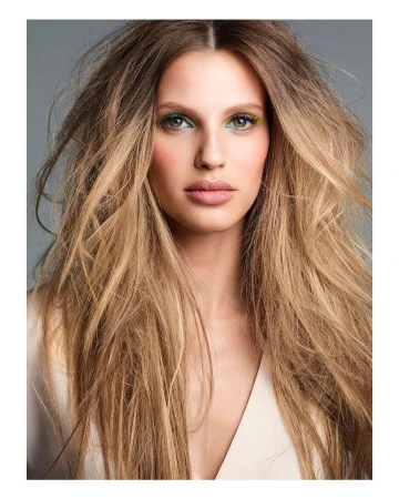 Close-up shot of a woman with long blonde hair, light skin and green eye shadow, wearing a light colored top, against a grey background.