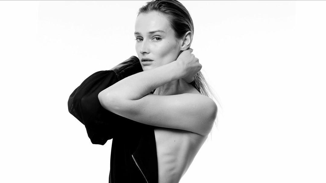 Black and white portrait of a woman with wet hair, holding a black leather jacket draped over her shoulders in a minimalist studio.