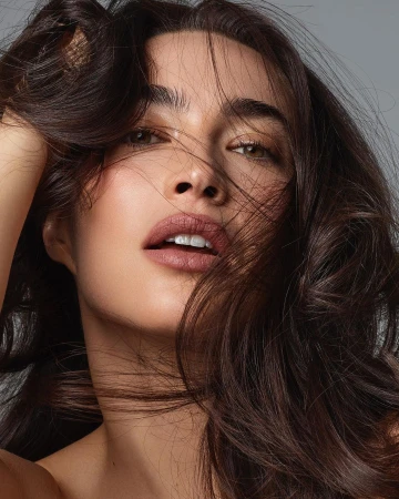 Close-up portrait of a brunette woman with hand in hair, showcasing soft skin tones, detailed hair texture, and a light gray background.