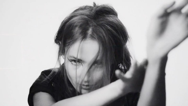 Close-up portrait of a woman with dark hair covering her face and dramatic eyeliner, raising her hands against a plain white background.
