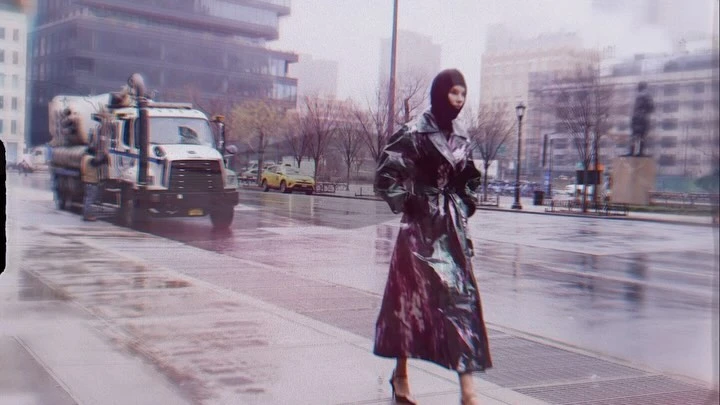 A woman in a shiny black coat walks on a wet city sidewalk, with a truck and buildings visible in the background on an overcast day.